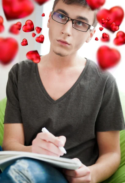Jonge volwassen man een Valentijn brief te schrijven aan zijn vriendin zittend op een grote Zitzak bij hem thuis. mooie rode harten zweven rond zijn hoofd — Stockfoto