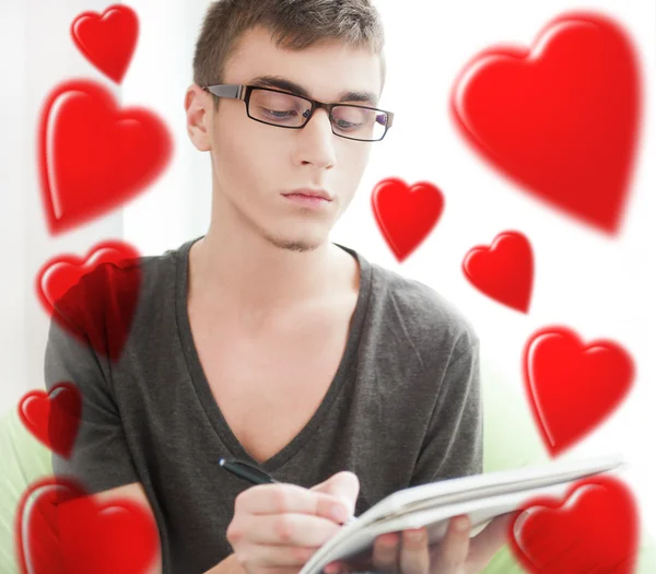 Portrait de jeune homme séduisant avec des cœurs volants autour de lui assis à sa maison et écrivant à l'aide de stylo et de copybook — Photo