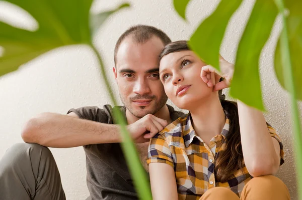 Attractive young adult couple sitting close on hardwood floor in home smiling and laughing. Monstera plant on foreground in blur — Stock Photo, Image