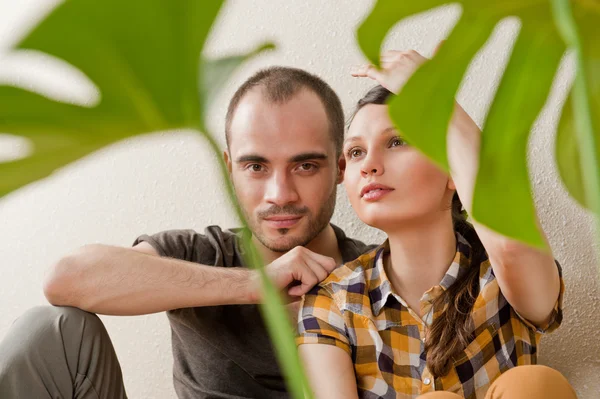 Atraente jovem casal adulto sentado perto do chão de madeira em casa sorrindo e rindo. Monstera planta em primeiro plano em borrão — Fotografia de Stock