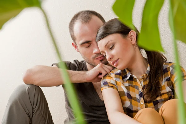 Attractive young adult couple sitting close on hardwood floor in home smiling and laughing. Monstera plant on foreground in blur — Zdjęcie stockowe