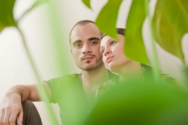 Attractive young adult couple sitting close on hardwood floor in home smiling and laughing. Monstera plant on foreground in blur — Zdjęcie stockowe