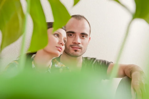Attractive young adult couple sitting close on hardwood floor in home smiling and laughing. Monstera plant on foreground in blur — Stock Photo, Image
