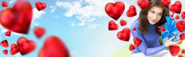 Mulher encantada abrindo um presente sentado no saco de feijão em casa e belas formas de coração vermelho estão voando ao redor, céu e nuvens no fundo — Fotografia de Stock