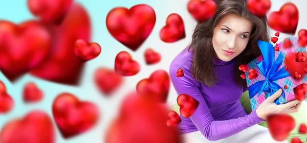 Delighted woman opening a gift sitting on the bean bag at home and beautiful red heart shapes are flying around. Valentine day concept poster — Stock Photo, Image