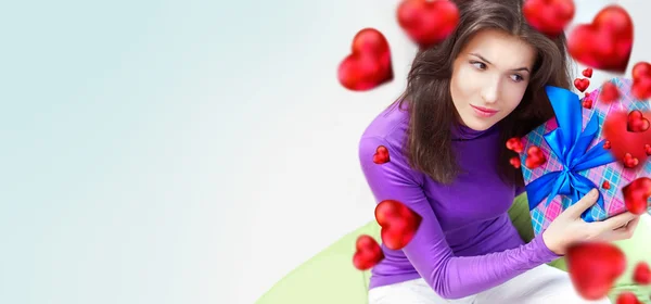 Delighted woman opening a gift sitting on the bean bag at home and beautiful red heart shapes are flying around. Valentine day concept poster — Stock Photo, Image