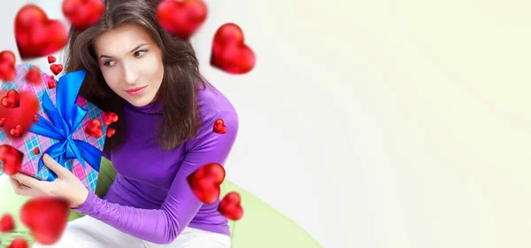 Delighted woman opening a gift sitting on the bean bag at home and beautiful red heart shapes are flying around. Valentine day concept poster — Stock Photo, Image