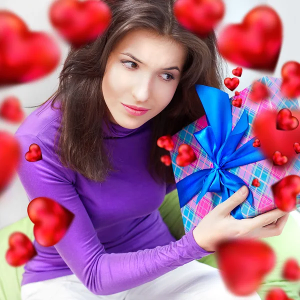 Glad kvinna öppnar en gåva som sitter på böna väska hemma och vackra röda hjärtat former flyger runt. Alla hjärtans dag konceptet affisch — Stockfoto