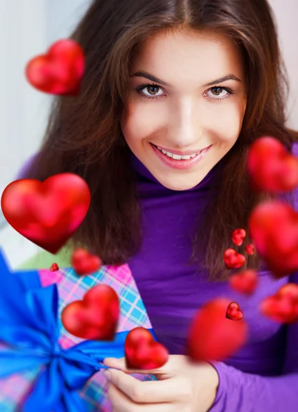 Задоволена жінка відкриває подарунок, сидячи на бобовій сумці вдома і красиві червоні форми серця літають навколо. День святого Валентина концепт плакат — стокове фото