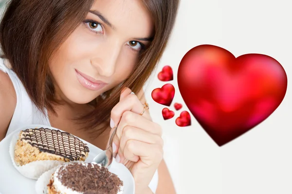 Smiling brunette woman eating some cake in the living room in her apartment. Graphic 3d red heart symbols are flying near her. She is in love — Stock Photo, Image