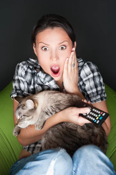 Jonge grappige vrouw zitten in haar zitzak en omarmen haar kat terwijl u tv kijkt. Ze is erg bang tijdens het kijken naar horrorfilm in donkere kamer — Stockfoto