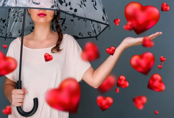 Giovane donna alla moda che tiene l'ombrello in piedi contro sfondo grigio cuori rossi galleggiano intorno a lei. Amore pioggia concetto — Foto Stock