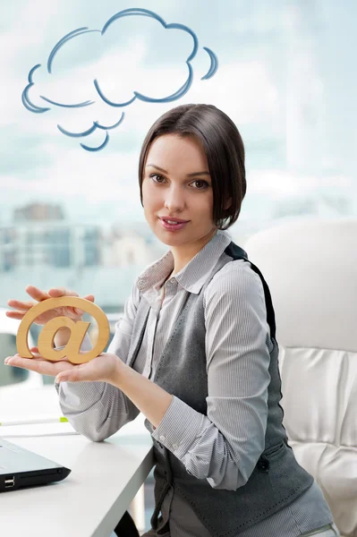 Portrait of a cheerful Business woman sitting on her desk holding an at symbol. Blank cloud balloon overhead — Stock Photo, Image