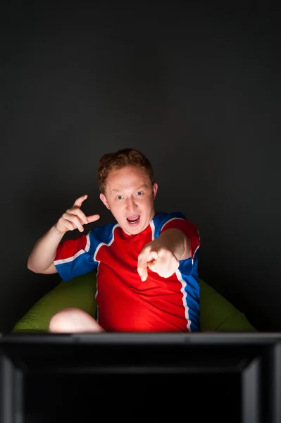 Close-up portrait of young man wearing sportswear fan of football team is watching tv and rooting for his favorite team. Sitting on beanbag alone at night. Light from television — Stock Photo, Image