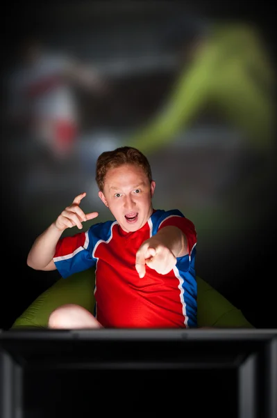Detailní portrét mladého muže nosit sportovní fanoušek fotbalové reprezentace je sledování televize a pro jeho oblíbeného týmu. sedí na beanbag sám v noci. světlo z televize — Stock fotografie