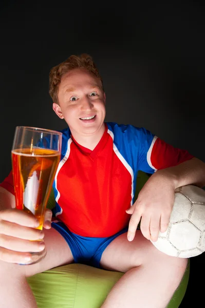 Νεαρός άνδρας κρατά μπάλα ποδοσφαίρου και μπύρα και βλέποντας τηλεόραση τη μετάφραση του παιχνιδιού στο σπίτι φορώντας αθλητικά είδη — Φωτογραφία Αρχείου