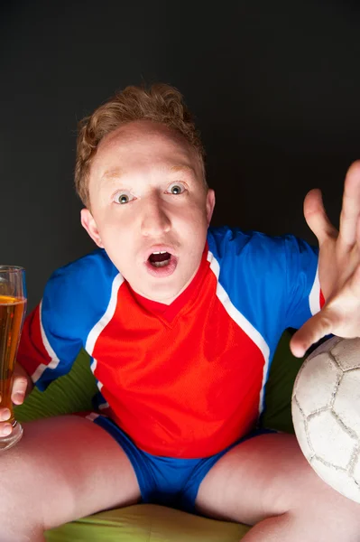 Молодий чоловік тримає футбольний м'яч і пиво і дивиться телевізійний переклад гри вдома в спортивному одязі — стокове фото