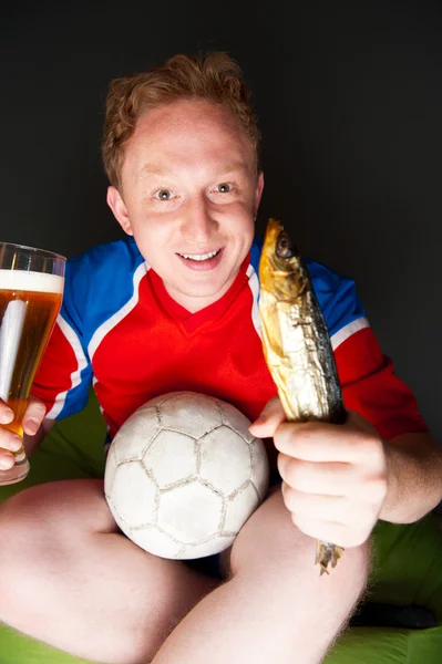 Young man holding soccer ball, beer and stockfish, watching tv translating of game at home wearing sportswear — Stock Photo, Image