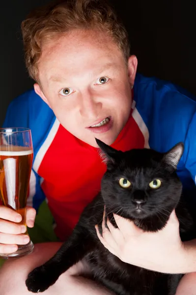Retrato de um jovem segurando um copo de cerveja e um grande gato preto e ambos olhando para a câmera enquanto assiste TV tradução de seu time de futebol favorito — Fotografia de Stock