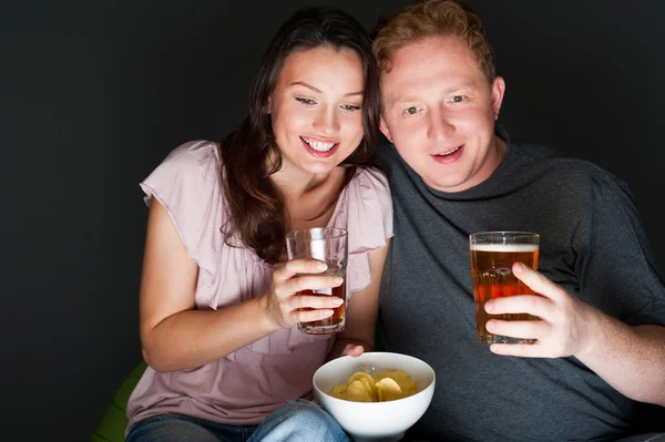 Gelukkige paar zit te samen kijken iets interessants op televisie drinken en eten - grijze achtergrond — Stockfoto