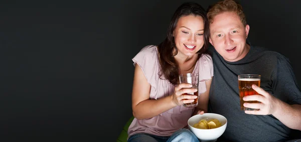 Glückliches Paar, das zusammen sitzt und im Fernsehen etwas Interessantes trinkt und isst - grauer Hintergrund — Stockfoto