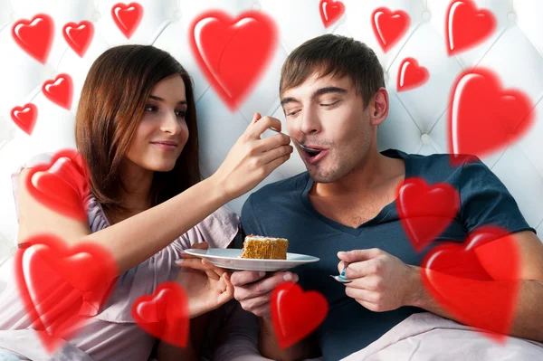 Portrait of happy couple lying in the bed. Girl feeding her boyfriend with spoon of tasty cake. Beautiful graphic red hearts are flying around — Stock Photo, Image