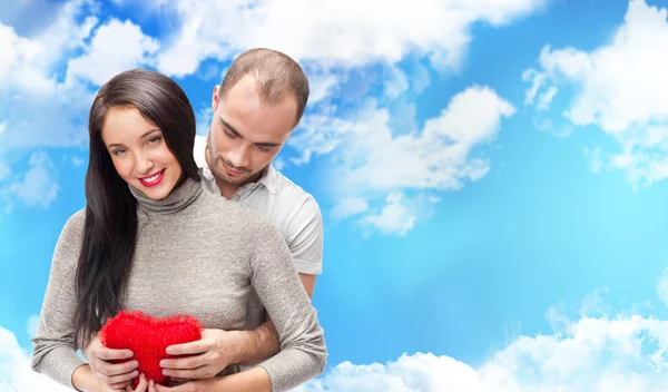 Feliz jovem casal adulto com coração vermelho no fundo romântico com céu e nuvens, abraçando e rindo — Fotografia de Stock