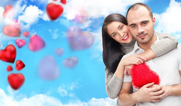 Happy young adult couple with red heart on romantic background with sky and clouds, embracing and laughing — Stock Photo, Image