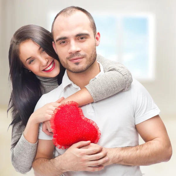 Retrato de jovem casal com coração vermelho em pé em sua casa e abraçando — Fotografia de Stock