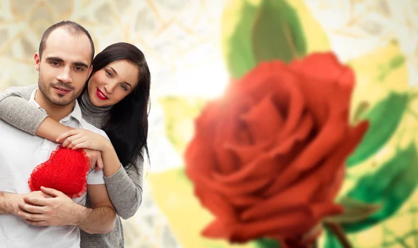 Glückliches junges erwachsenes Paar mit rotem Herz auf Hintergrund mit roter Rosenknospe, umarmt und lacht — Stockfoto