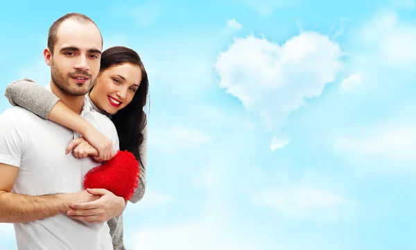 快乐年轻成人夫妇与浪漫背景与天空和云层，拥抱着，笑着上红色的心 — 图库照片