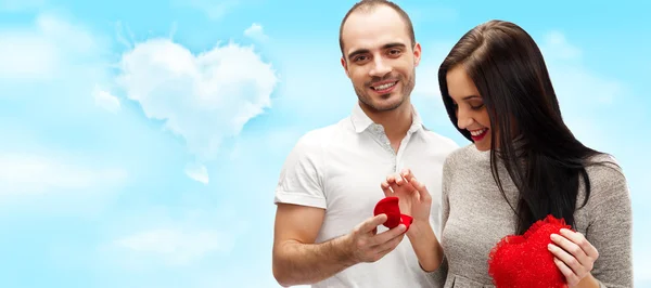 Boldog fiatalember gifting egy gyűrűt, hogy egy gyönyörű meglepett fiatal nő romantikus háttér a sky és a szív alakú felhők — Stock Fotó