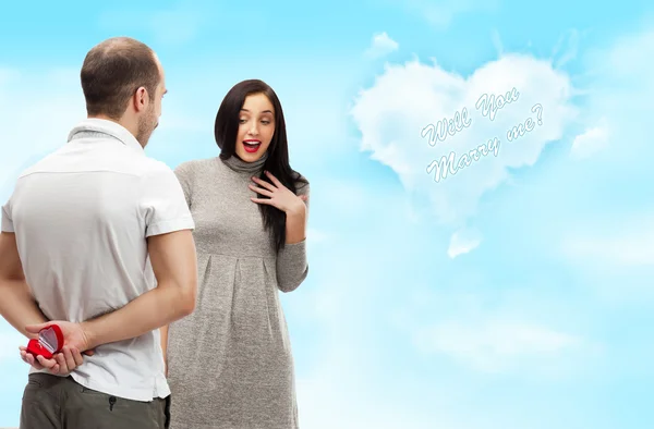 Happy young man gifting a ring to a beautiful surprised young woman on romantic background with sky and clouds of heart shape — Stock Photo, Image
