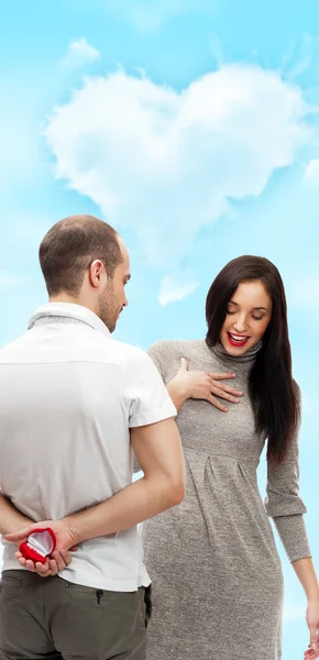 Glücklicher junger Mann, der einer schönen überraschten jungen Frau auf romantischem Hintergrund mit Himmel und Wolken in Herzform einen Ring schenkt — Stockfoto