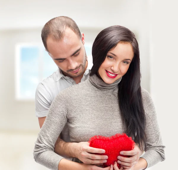 Retrato de jovem casal com coração vermelho em pé em sua casa e abraçando — Fotografia de Stock