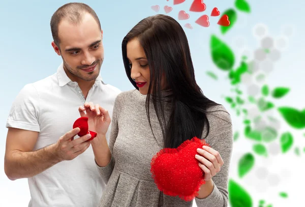 Proposal of marriage: young man putting ring on young ladys finger, on romantic background with red hearts and green leaves Stock Image