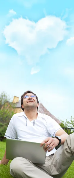 Een glimlachende man met laptop buiten zittend in zijn achtertuin en opkijkend naar de wolk van hartvorm boven zijn hoofd — Stockfoto
