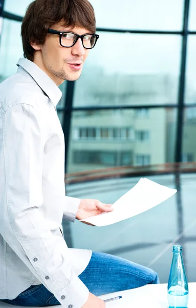 Close-up portrait of young creative businessman looking at camera while sitting relaxed on table at his office and holding papers — Stok fotoğraf