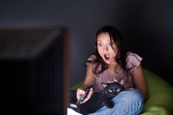 Joyeux jeune femme échangeant les chaînes de télévision, détendue sur le canapé du sac à haricots. Gros chat noir couché avec elle et dormant . — Photo