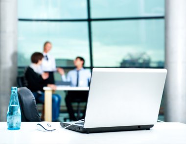 Modern laptop on foreground at empty workplace. Team of young businesspeople discussing plans on background clipart