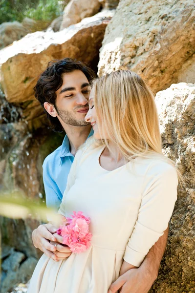 Retrato de pareja de amor abrazando al aire libre en el parque mirando feliz. Mujer sosteniendo flor. Cascada en el fondo —  Fotos de Stock