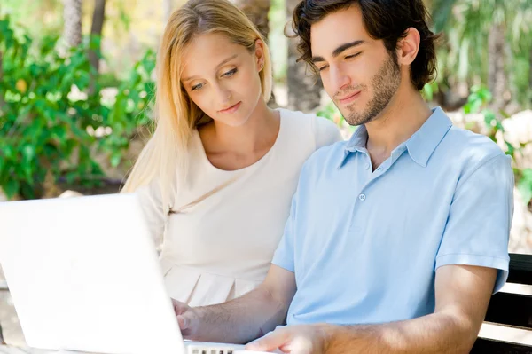 Jovem casal trabalhando no laptop e sorrindo enquanto sentado relaxado no banco no parque de verão — Fotografia de Stock