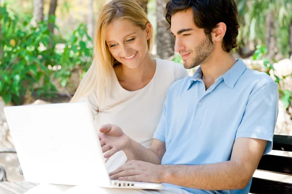 Молода пара працює на ноутбуці і посміхається, сидячи розслабленим на лавці в літньому парку — стокове фото