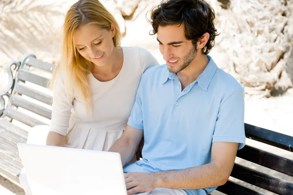 Dolgozik a laptop, és mosolyogva, miközben ül a fiatal pár nyugodt nyári parkban a padon — Stock Fotó