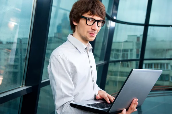 Χαρούμενος νεαρός επιχειρηματίας χρησιμοποιώντας φορητό υπολογιστή στην επιχείρηση κτίριο, χαμογελώντας. — Φωτογραφία Αρχείου