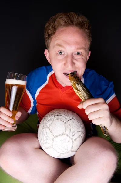Portret młodzieńca, oglądanie tv tłumaczenie o meczu piłki nożnej z jego ulubionej drużyny i picia piwa i jedzenia słonych ryb — Zdjęcie stockowe