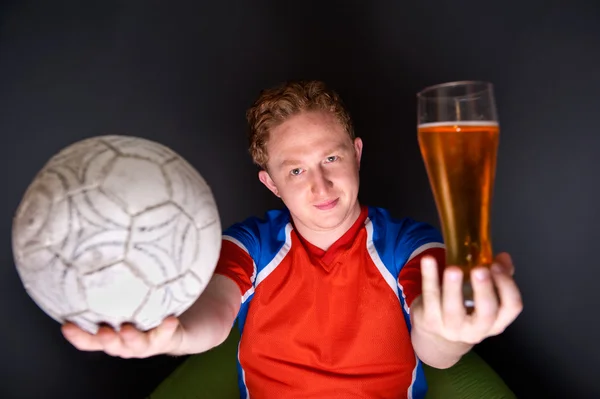 Портрет молодого чоловіка, який дивиться телепереклад футбольного матчу зі своєю улюбленою командою і тримає м'яч і п'є пиво — стокове фото