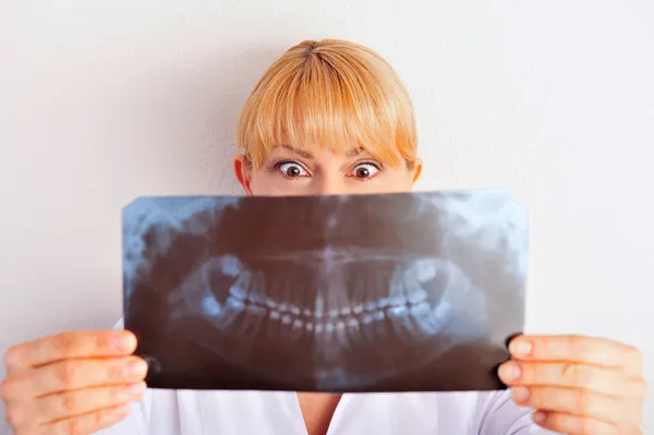 Retrato de una graciosa doctora caucásica sonriente vistiendo uniforme de pie contra la pared en el hospital mirando los resultados de rayos X de su paciente y ella está muy sorprendida —  Fotos de Stock
