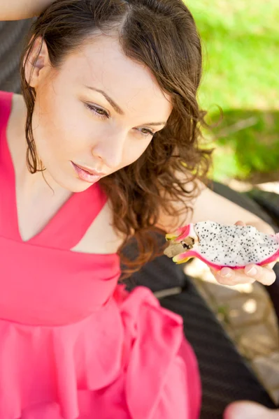 Retrato de una joven bonita mujer con vestido rosa brillante comiendo frutas exóticas de dragón asiático y disfrutando de sus vacaciones en un resort tropical —  Fotos de Stock