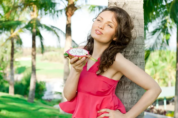 Portré, fiatal, csinos nő visel világos rózsaszín ruha eszik egzotikus ázsiai sárkány gyümölcs, és élvezi vele nyaralni: tropical resort — Stock Fotó